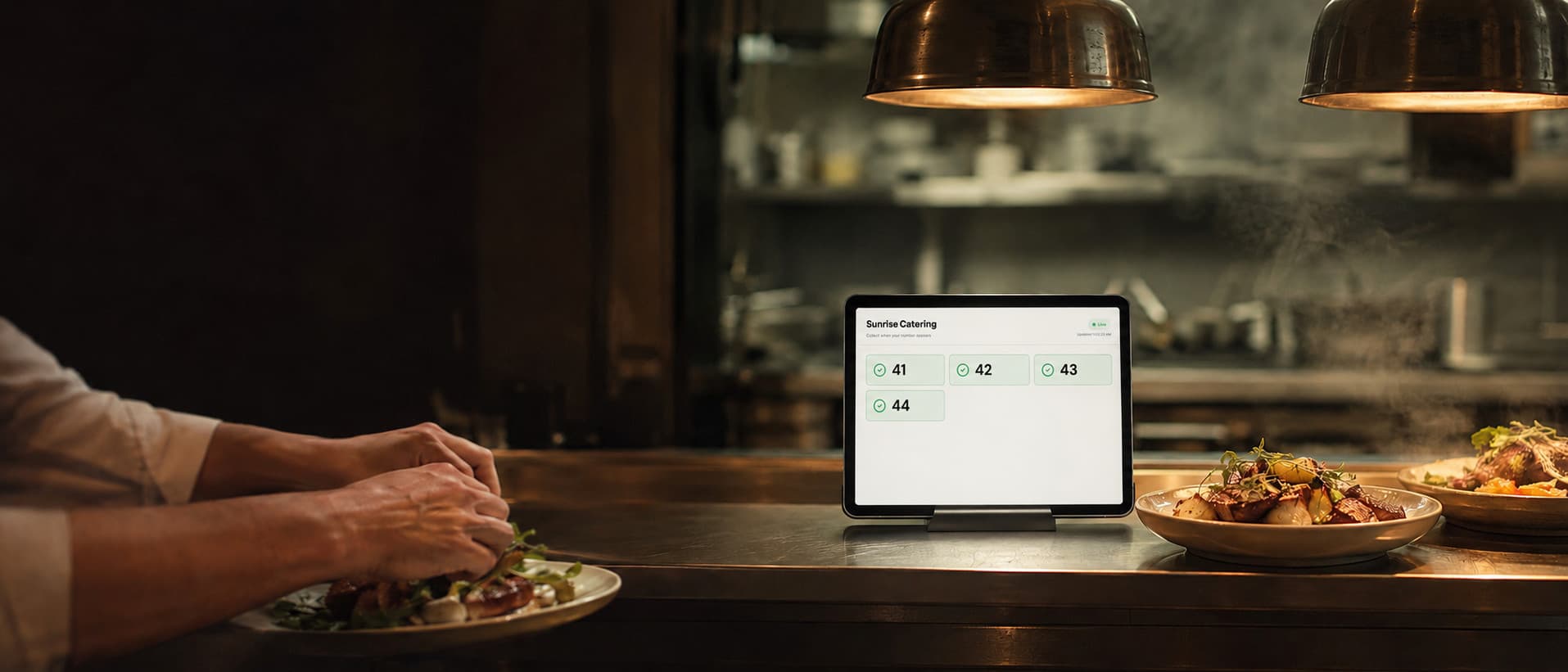 A working kitchen pass under warm heat lamps; where a terminal would stand there is an ordinary tablet in a simple stand.